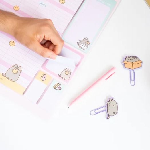 Ръката откъсва бележка от 16-месечния блок за планиране на Grupo Erik Pusheen The Cat (септември 2025 г. - декември 2026 г.), заобиколена от розова химикалка и кламери с котешки мотиви - идеални за освежаване на организационния календар или планер с аксес-4