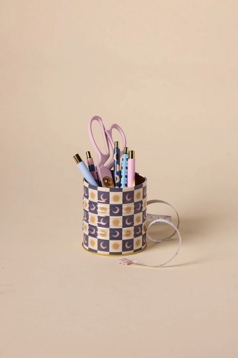 Шарената чаша от DesignWorks Ink със сини и златни мотиви на слънце и луна държи пастелни химикалки и ножици с розови дръжки. До метален моливник на бежов фон е навита бяла измервателна лента.-2