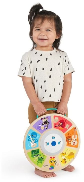 Малко дете с тъмна коса, облечено в бяла риза на черни точки и кафяви панталони, се усмихва, докато държи Музикална играчка Hape Baby Einstein, сензорна - цветна и забавна за сетивна игра.-3