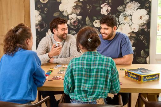 Четирима души играят на настолната игра Splendor, обновено издание, усмихнати и ангажирани в търговия със скъпоценни камъни с карти и цветни жетони. На заден план има флорални тапети и слънчева светлина от прозореца.-4