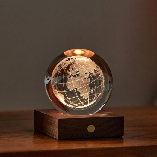 Нощна лампа Gingko Amber Crystal Light World Globe представлява стъклен глобус с гравирана карта на света, осветен отгоре, поставен върху лампа орехово дърво със златна точка върху дървена повърхност на тъмен фон.-3