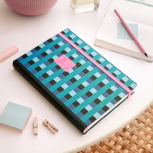 Тефтерът - органайзер Miquelrius Grafica Color Block 2026, ден на страница със синя, зелена и черна карирана корица и розов ластик е на кръгла бяла маса сред тефтер, розов молив, самозалепващи се листчета и щипки.-8