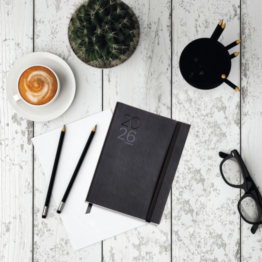 Плосък план на 2026 тефтер-органайзер Miquelrius Luxor & Ágora Negro, ден на страница, с кожени корици, два молива, празна хартия, кафе, очила, кактус и черен моливник върху бяла селска маса.-6