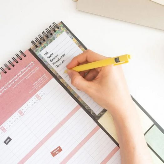 Ръка с жълта химикалка пише списък с хранителни продукти (мляко, ябълки, банани, минерална вода, сирене, шоколад) върху тефтерче в горната част на планер-пад Grupo Erik Van Gogh за 16 месеца - септември 2025 / декември 2026 година на бяло бюро.-3