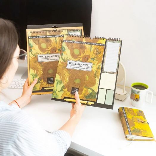Човек на бюро държи слънчогледова тематика Планер-пад Grupo Erik Van Gogh за 16 месеца (септември 2025 - декември 2026 г.), с подходящи бележник, стикери за планер, чаша и тетрадка до компютърен монитор.-6