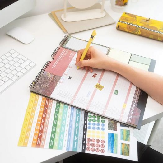 Някой пише в спирала Grupo Erik Van Gogh планер-пад за 16 месеца (септември 2025 - декември 2026) на бюро с цветни стикери за планер и клавиатура наблизо, използвайки жълт молив.-4