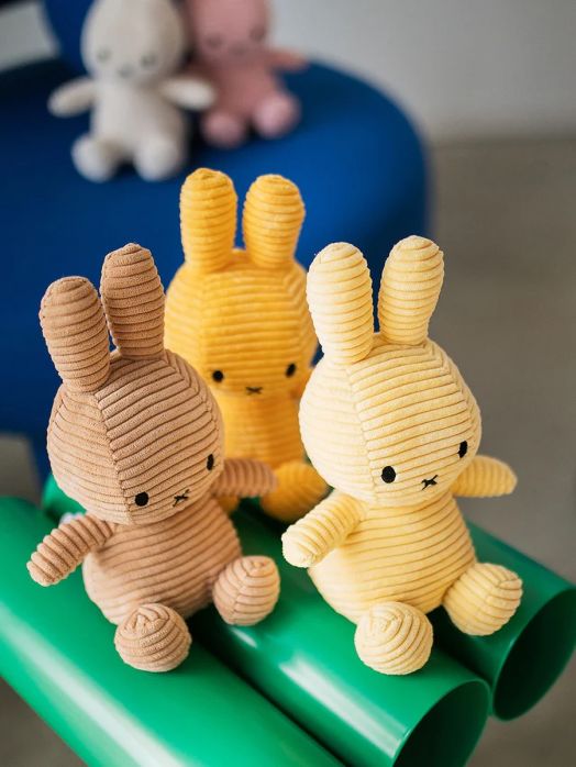 Кремаво-жълто плюшено зайче Miffy Eco Corduroy (23 cm) седи върху яркозелена тръба, а на заден план се виждат още две размазани плюшени зайчета върху синя повърхност.-4