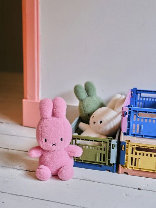 A плюшена играчка Miffy Eco Corduroy - розов заек, 23 см седи върху бял под до три цветни щайги с още два плюшени заека - зелен и бял, край стена с розова каса на врата.-4