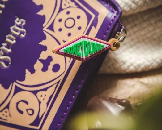 Близък план на лилаво-синьо с цип Портмоне ABYstyle Хари Потър - Шоколадово жабче с прикрепен зелен и розов етикет Honeydukes, до парче бонбони Хари Потър Шоколадово жабче върху текстурирана кремава повърхност.-4