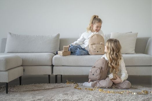Две малки момичета с ранички за детска градина „Vadobag Kidzroom Comfort Friends“ с мотиви на животни седят на бежов диван и килим и се усмихват една на друга. Едното момиче държи опаковани подаръци, а по килима блестят лампички. Атмосферата е уютна и изп-7