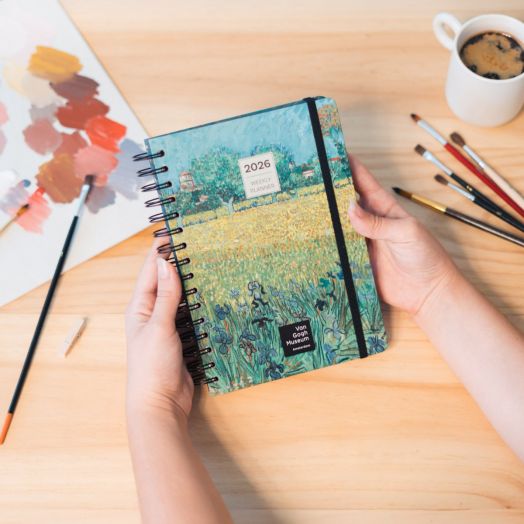 Някой държи седмичен тефтер-органайзер Grupo Erik Van Gogh 2026, а на дървената маса на заден план седят четки за рисуване, палитра и чаша кафе.-3