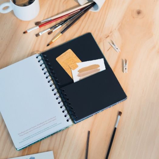 Седмичен тефтер-органайзер Grupo Erik Van Gogh 2026 в спираловиден формат А5 е отворен на дървена маса с карти в черен джоб, заобиколен от четки за рисуване, чаша за кафе и щипки за дрехи - идеален за планиране на седмицата през 2026 г.-7