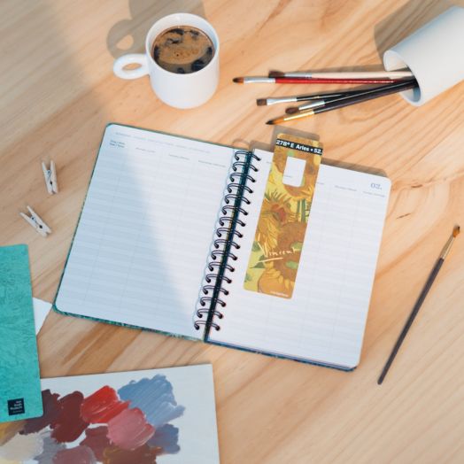 Открит Седмичен тефтер-органайзер Grupo Erik Van Gogh 2026 лежи на дървено бюро с разделител, чаша кафе, четки за рисуване, преобърната чаша, малки дървени щипки и цветна палитра.-4