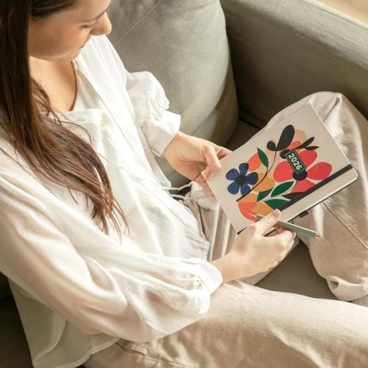 Жена с бяла риза седи на диван, държи цветна Седмичен тефтер - Оргаизер Miquelrius Natura Mini Garden 2026 и молив, като се готви да пише или да си прави бележки в нея.-8