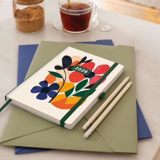 Седмичен тефтер-органайзер Miquelrius Natura Mini Garden 2026 с флорална корица лежи върху зелени и сини папки, а на масата има два бели молива, чаша чай с лъжица и буркан кафява захар.-7