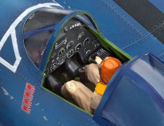 Близък план на пилотската кабина от Сглобяем модел Revell - Самолет F4U-1A Corsair, включващ фигура на пилот с оранжева каска, бежова униформа, жълта жилетка и детайлни сини прибори и контролни уреди.-5