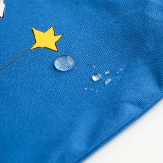 Водни капки се стичат по синята материя на шопинг чанта Grupo Erik - Малкият принц в близост до жълта звезда, подчертавайки водоотблъскващото ѝ свойство.-3