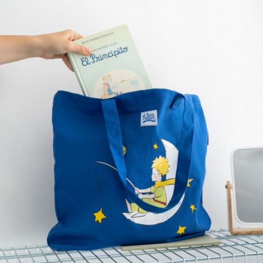 Ръка поставя испанско издание на "Малкият принц" в синя кутия Grupo Erik - The Little Prince шопинг чанта с героя върху полумесец, поставена върху бяла повърхност до малко огледало.-6