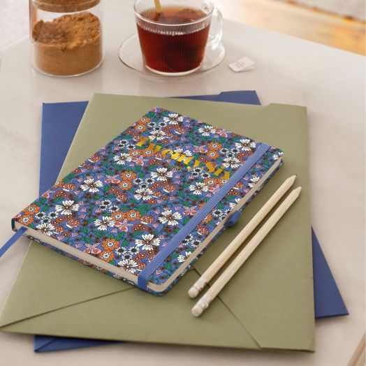 A Тефтер - органайзер Miquelrius Natura Blooming 2026, ден на страница, с флорални мотиви и син ластик, лежи върху зелени и сини папки до два бели молива на маса с чаша чай, бурканче кафява захар и пакетче захар.-3