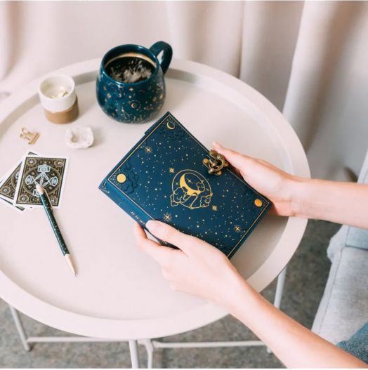 Човек държи тефтер с корици от веган кожа Grupo Erik - Astral в тъмносиньо с астрологични мотиви и златни звезди, разположен на бял кръг плот сред чаша, химикал, карти и кристали.-7