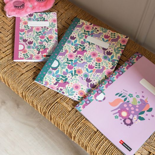 Три тетрадки Miquelrius Flower Unicorn A5, с 48 широки реда, украсени с еднорози и флорални мотиви в лилаво, розово и зелено, са подредени върху плетена повърхност до розова пухкава маска за сън.-2