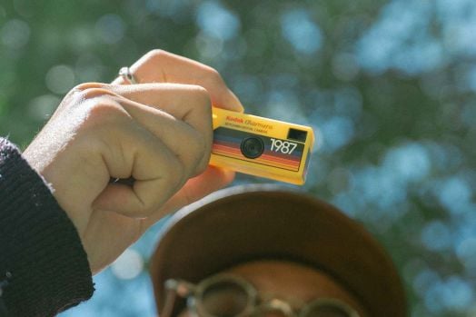 Човек държи ретро стил фотоапарат - по-специално компактен жълт Цифров фотоапарат Kodak - Charmera Keychain, асортимент. Фотоапаратът е с ясен фокус, докато лицето на човека и фонът от листа са размазани.-12