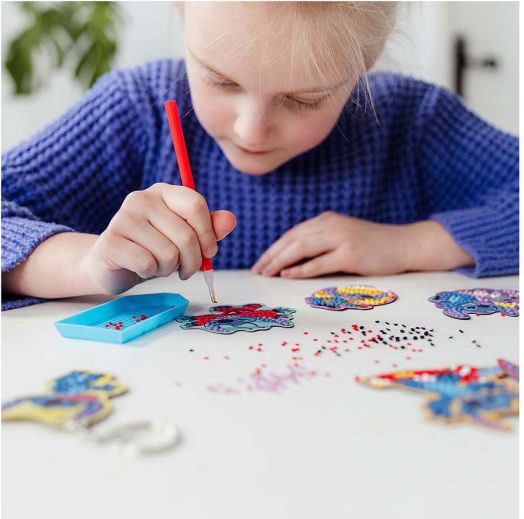 Дете в син пуловер използва Творчески комплект Totum Stitch 3 в 1, за да украси цветни пеперуди с червени инструменти върху бяла маса, заобиколена от малки мъниста и гривни за направа.-4