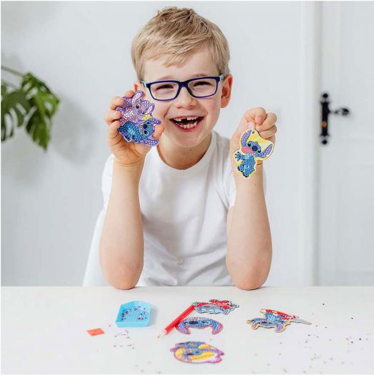 Усмихнато момче с очила държи две цветни животни от Творчески комплект Totum Stitch 3 в 1, седнало на бяла маса с още принадлежности за занаяти и декорации пред него.-5