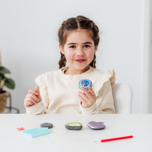 Младо момиче със сплетена коса седи на бяла маса, усмихва се и държи изрисуван камък от Творчески комплект Totum Stitch - Декориране на камъни, 4 броя. Други декоративни камъчета и принадлежности за изкуство са разпръснати на масата пред нея.-4