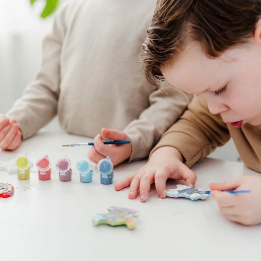 Две деца рисуват малки дървени фигури на бяла маса с пастелни бои - перфектно забавление с Творчески комплект Totum Stitch - Направи и оцвети гипсови фигури.-3