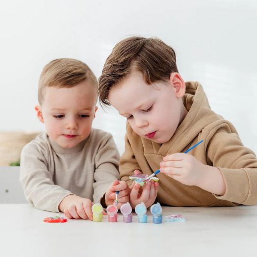 Две деца оцветяват гипсови фигурки Стич от Творчески комплект Totum Stitch - Направи и оцвети, едното рисува с четка, а другото гледа, заобиколени от цветни боички на масата.-4