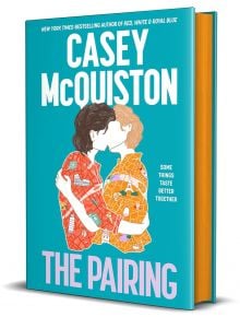 3D корицата на книгата The Pairing представя двама илюстрирани души - единият с тъмна коса, другият с руса - които се целуват на тиландски фон с удебелен бял и розов текст.